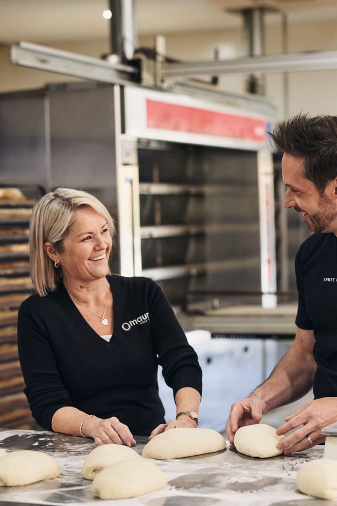 A man and a woman smiling at each other kneading dough in a bakery kitchen