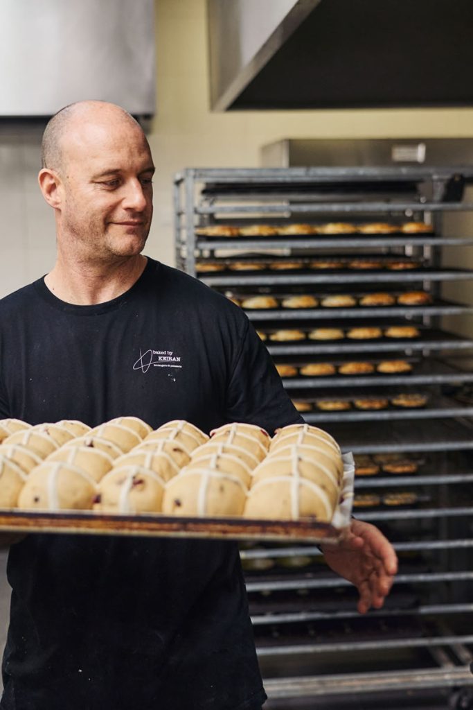 Male baker in a black t-shirt holcing a tray of hot cross buns