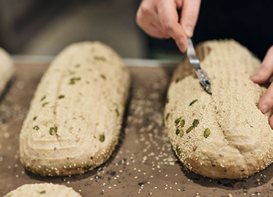 close-up image of a baker using a lame to score the top of a seeded loaf of unbaked bread