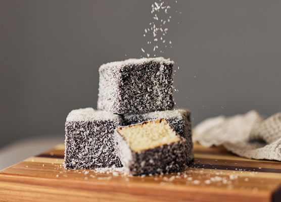 Baked lamingtons stacked on a wooden board with shredded coconut sprinkled over